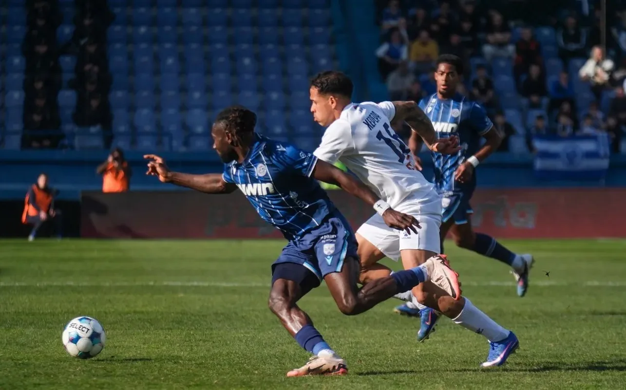 Dva fudbalera u akciji na terenu, jedan u plavom dresu pokušava oduzeti loptu igraču u bijelom dresu.