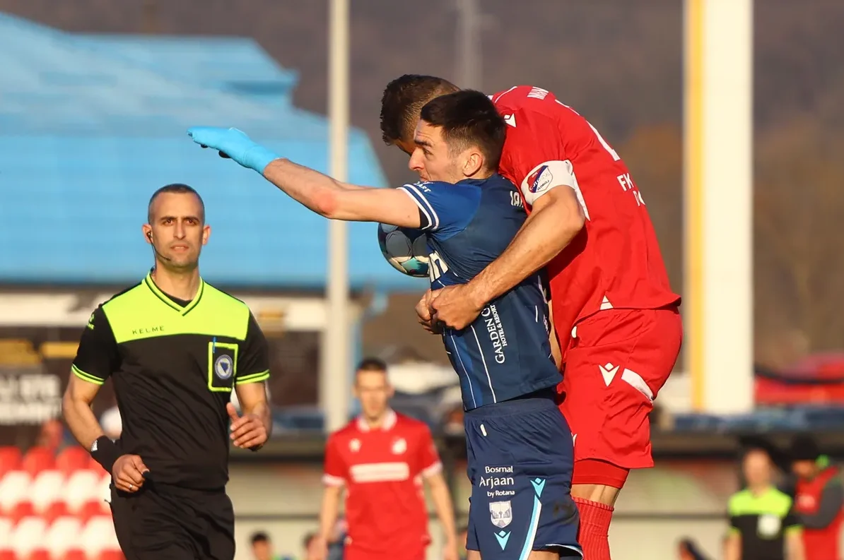 Fudbaler u plavom dresu drži loptu, dok ga igrač u crvenom dresu drži, a sudija sa strane pomno prati situaciju.