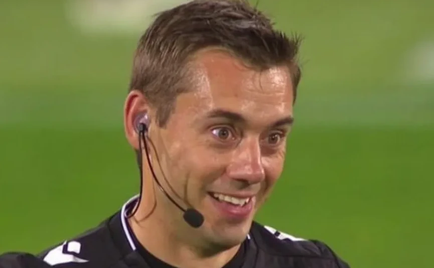 Fudbalski sudija sa slušalicama i mikrofonom, gleda u kameru sa osmijehom.