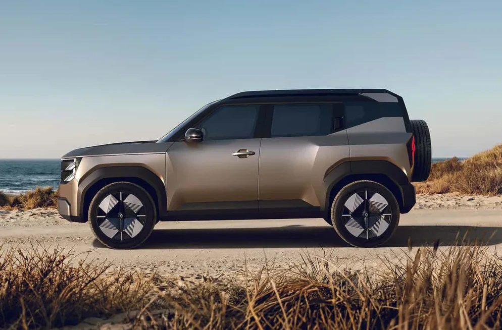 Futuristički koncept SUV vozila u boji šampanjca, sa crnim krovom i detaljima, parkiran na pješčanoj stazi pored mora. Vozilo ima moderno dizajnirane točkove i rezervni točak montiran na stražnjoj strani.