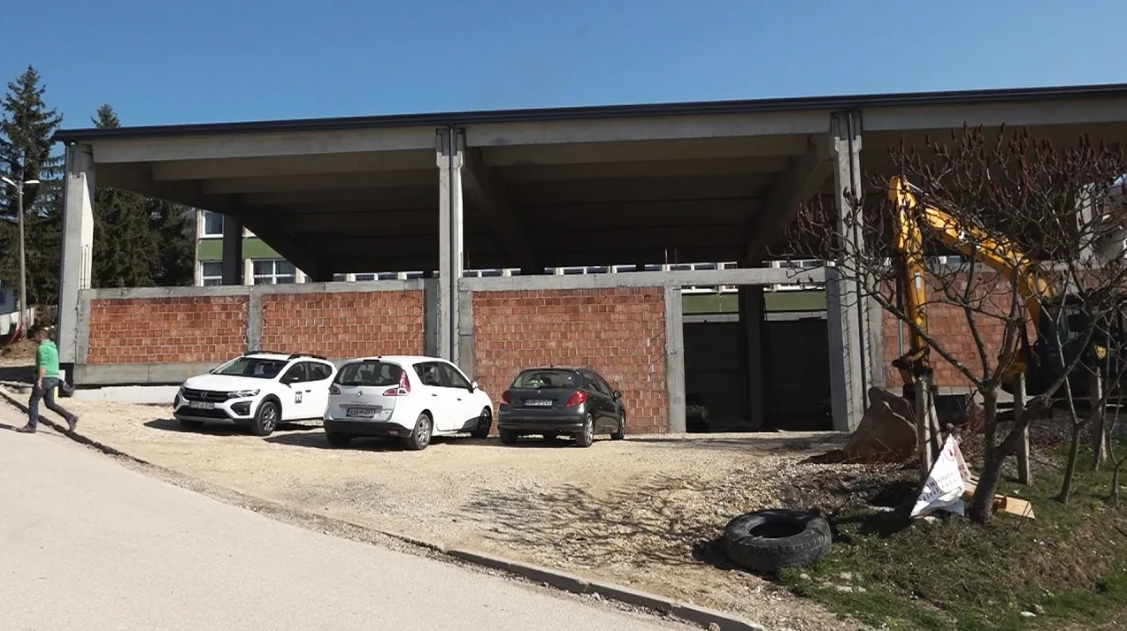 Gradilište u fazi izgradnje sa tri parkirana automobila, bagerom i ciglama.