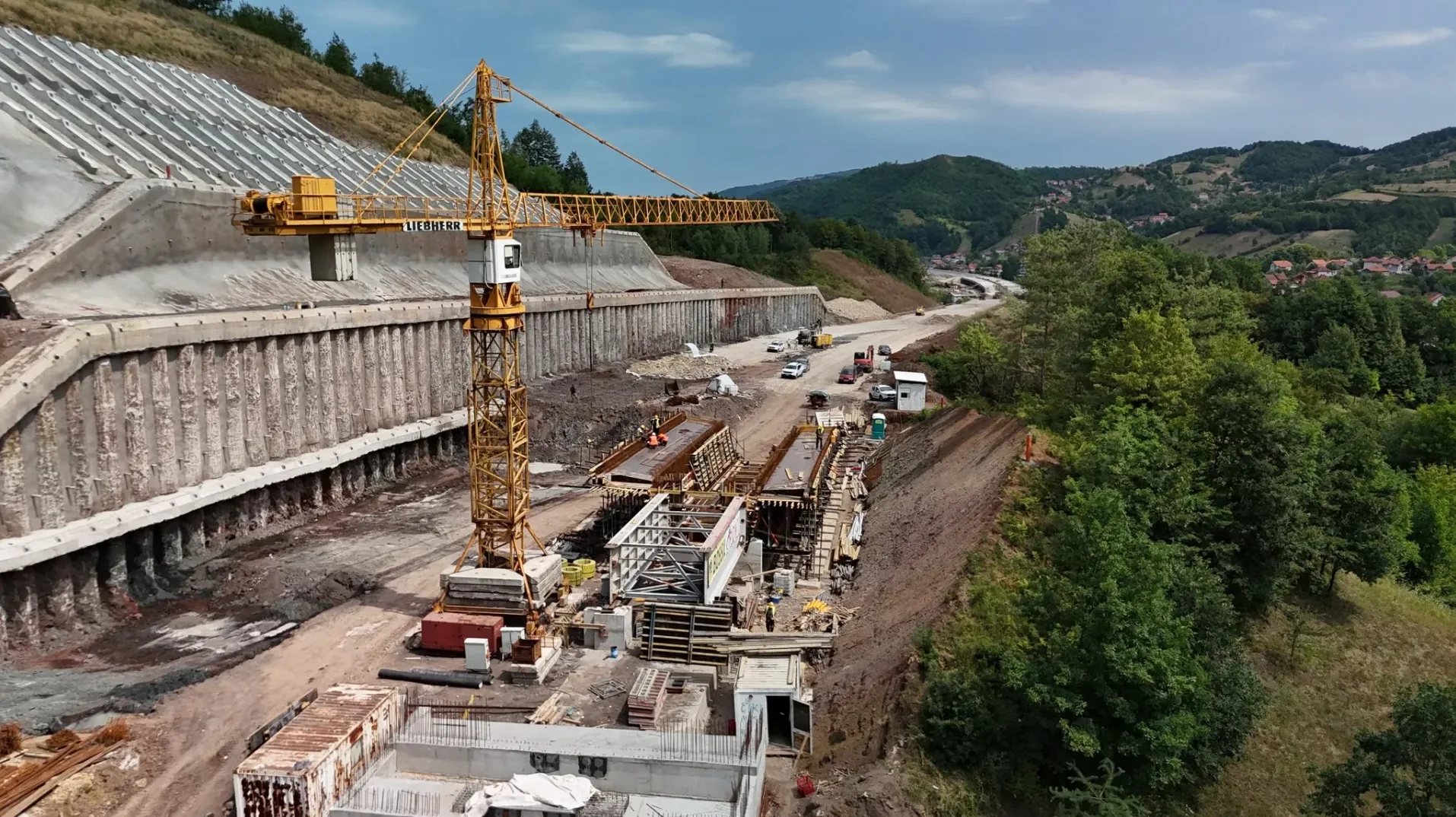Veliki žuti kran na gradilištu pored potpornog zida i započete izgradnje puta, s drvećem i kućama u pozadini.
