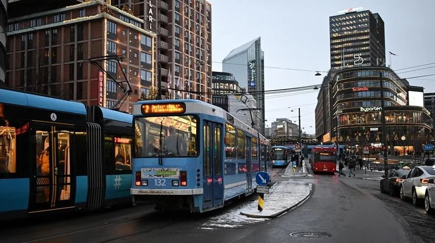 Plavi tramvaj broj 132 vozi ulicom u gradu, pored njega drugi tramvaj i crveni autobus, u pozadini moderne zgrade i ulična rasvjeta.