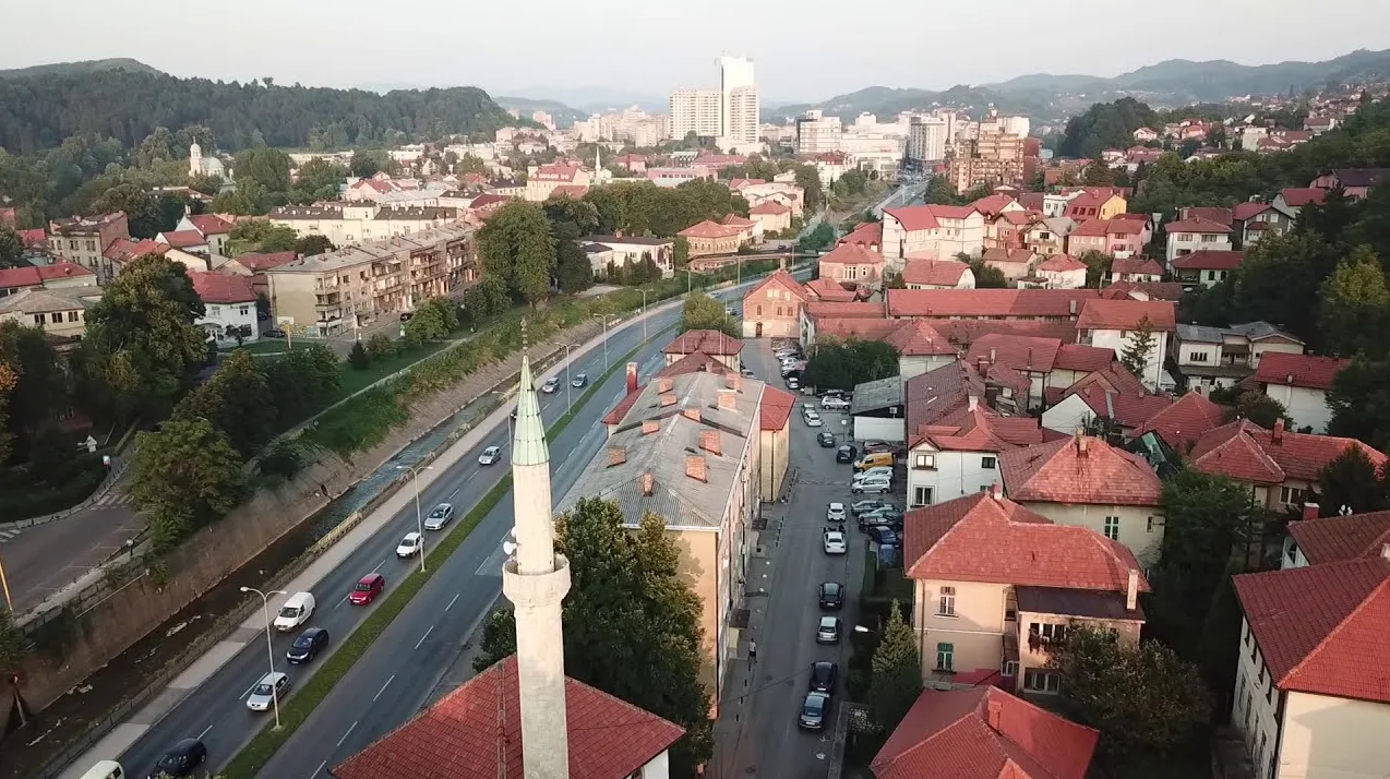 Avionski pogled na gradski pejzaž sa minaretom džamije u prvom planu, ulicom kojom saobraćaju automobili, stambenim zgradama sa crvenim krovovima i brdima u pozadini.