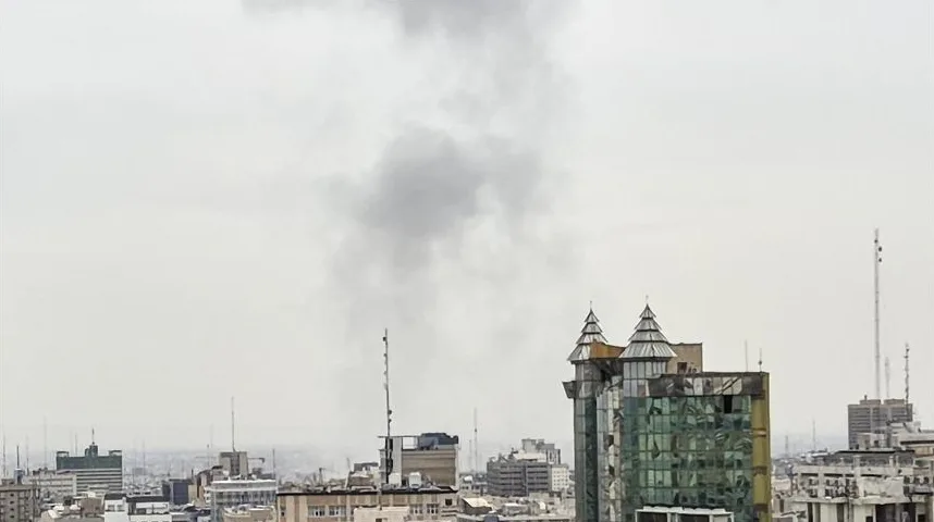 Dimni stub iznad urbanog gradskog pejzaža sa zgradama različitih visina i arhitektura, uključujući modernu zgradu sa staklenom fasadom i krovnim tornjićima.
