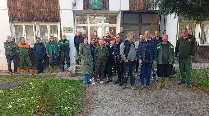 Grupa radnika u zelenim uniformama pozira ispred drvene zgrade.