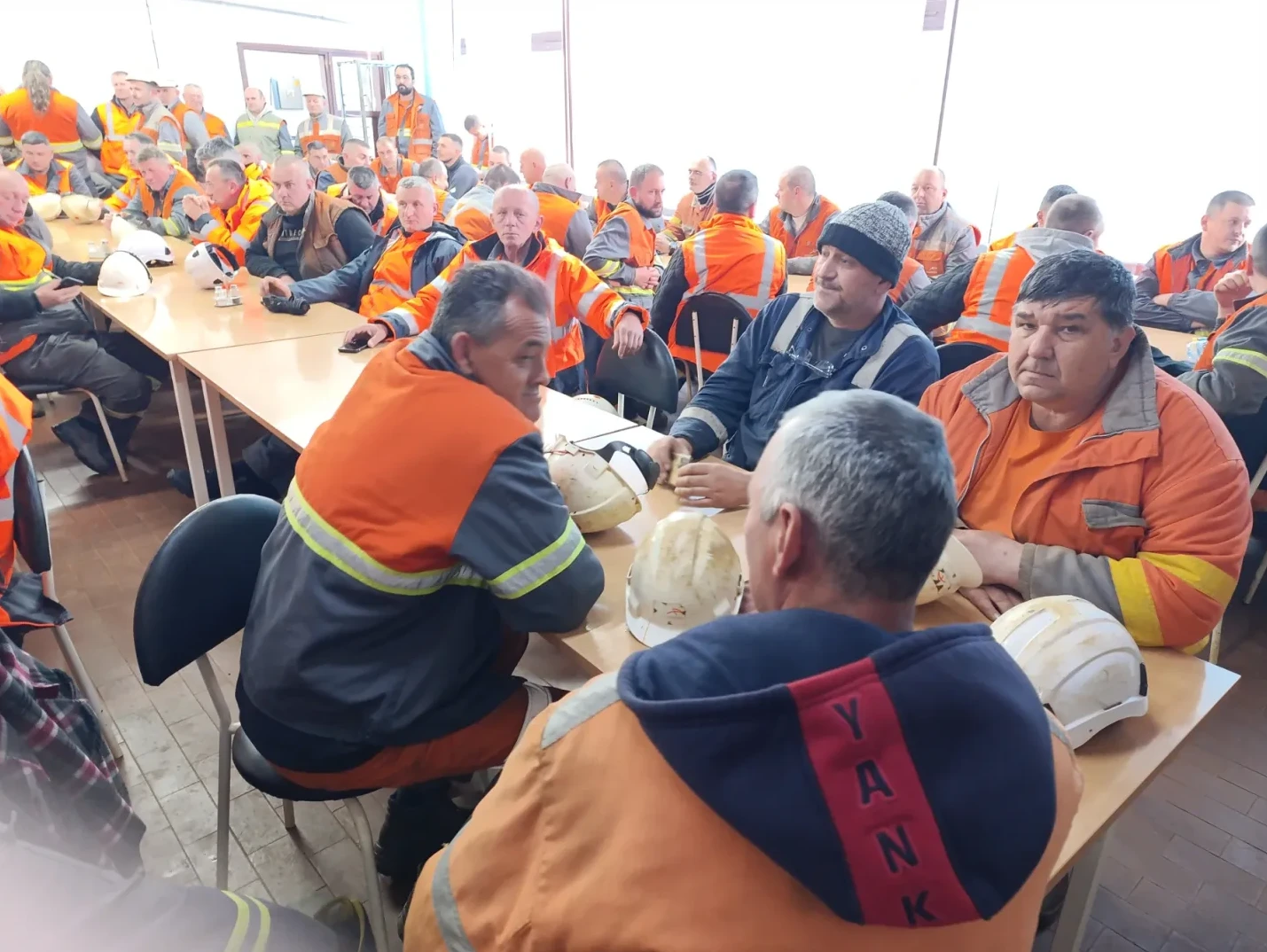 Grupa radnika u narandžastim radnim odijelima sjedi za stolovima na sastanku, sa zaštitnim kacigama na stolovima.