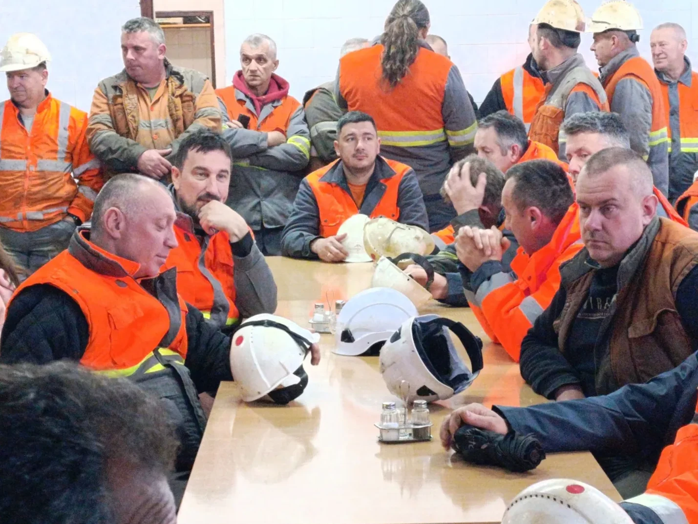 Grupa muškaraca u narandžastim prslucima i zaštitnim kacigama sjedi za stolom.