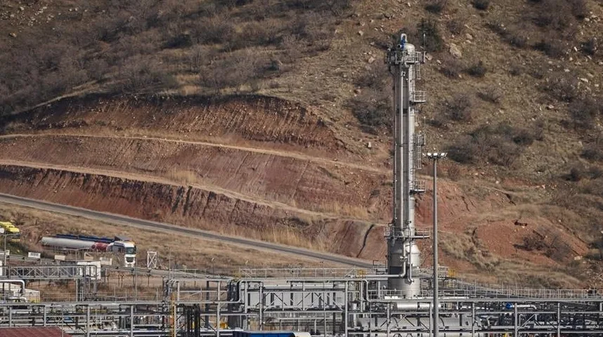 Industrijsko postrojenje s tornjem za destilaciju i cjevovodima, smješteno uz obronak brda obraslog suvom vegetacijom i stijenama.