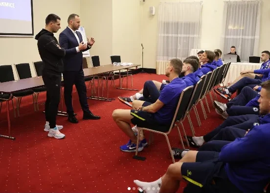 Trener ili vođa tima, obučen u tamno odijelo, drži predavanje grupi sportista obučenih u plave trenerke. Sportisti sjede u redovima na stolicama u sali sa crvenim tepihom.