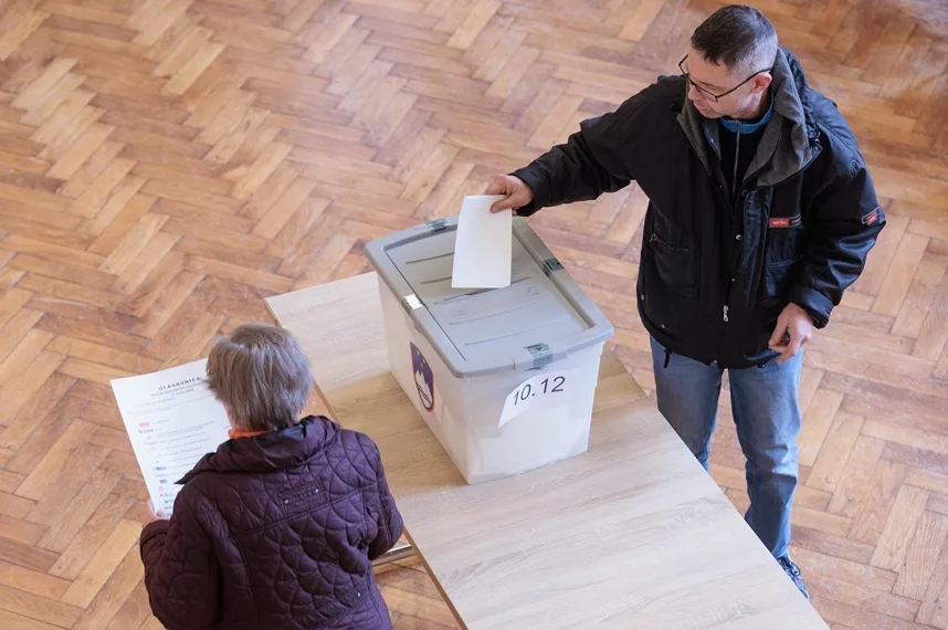 Čovjek ubacuje glasački listić u glasačku kutiju, dok žena stoji pored i drži glasački listić.