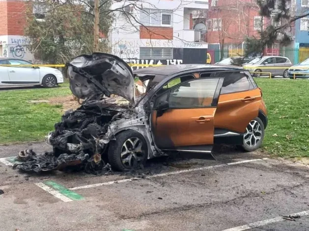 Narandžasti automobil uništen požarom na parkingu, sa otvorenim vratima i haubom.