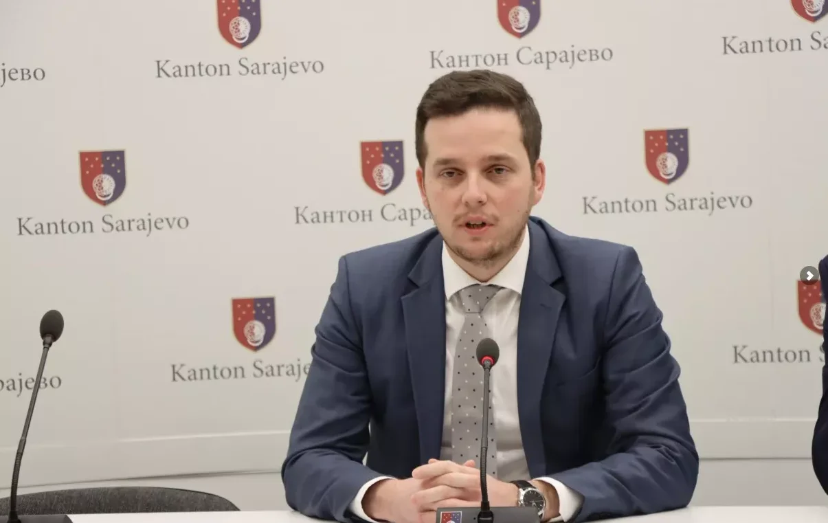 Čovjek u tamnom odijelu s bijelom košuljom i sivom kravatom sjedi za stolom ispred mikrofona. U pozadini se vidi grb Kantona Sarajevo.
