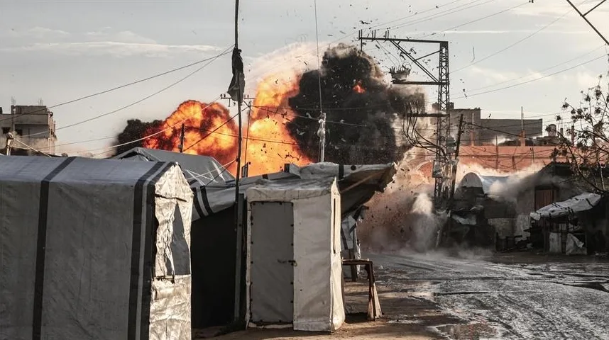 Moćna eksplozija u urbanom okruženju, s gustim dimom i plamenom koji izbija u blizini privremenih šatora i oštećenih zgrada.