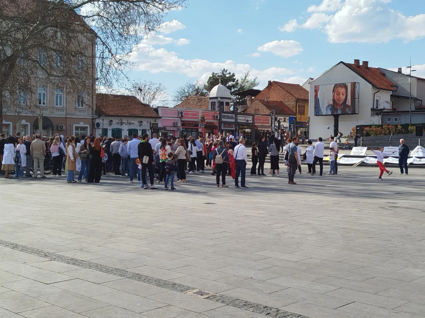 Grupa ljudi stoji na popločanom trgu ispred zgrada, dok se na velikom ekranu vidi projekcija mlade djevojke.