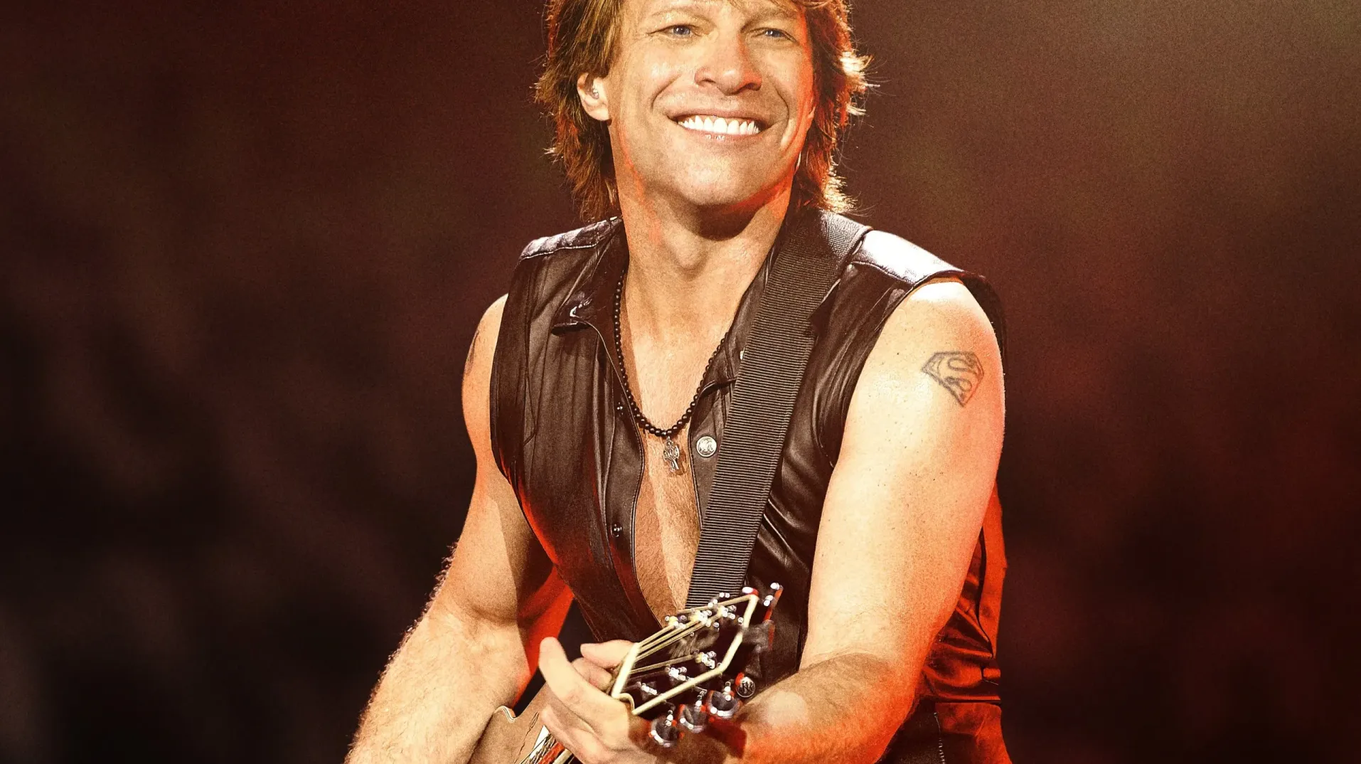 Džon Bon Džovi u kožnoj jakni bez rukava i s ogrlicom svira gitaru na osvijetljenoj pozornici.