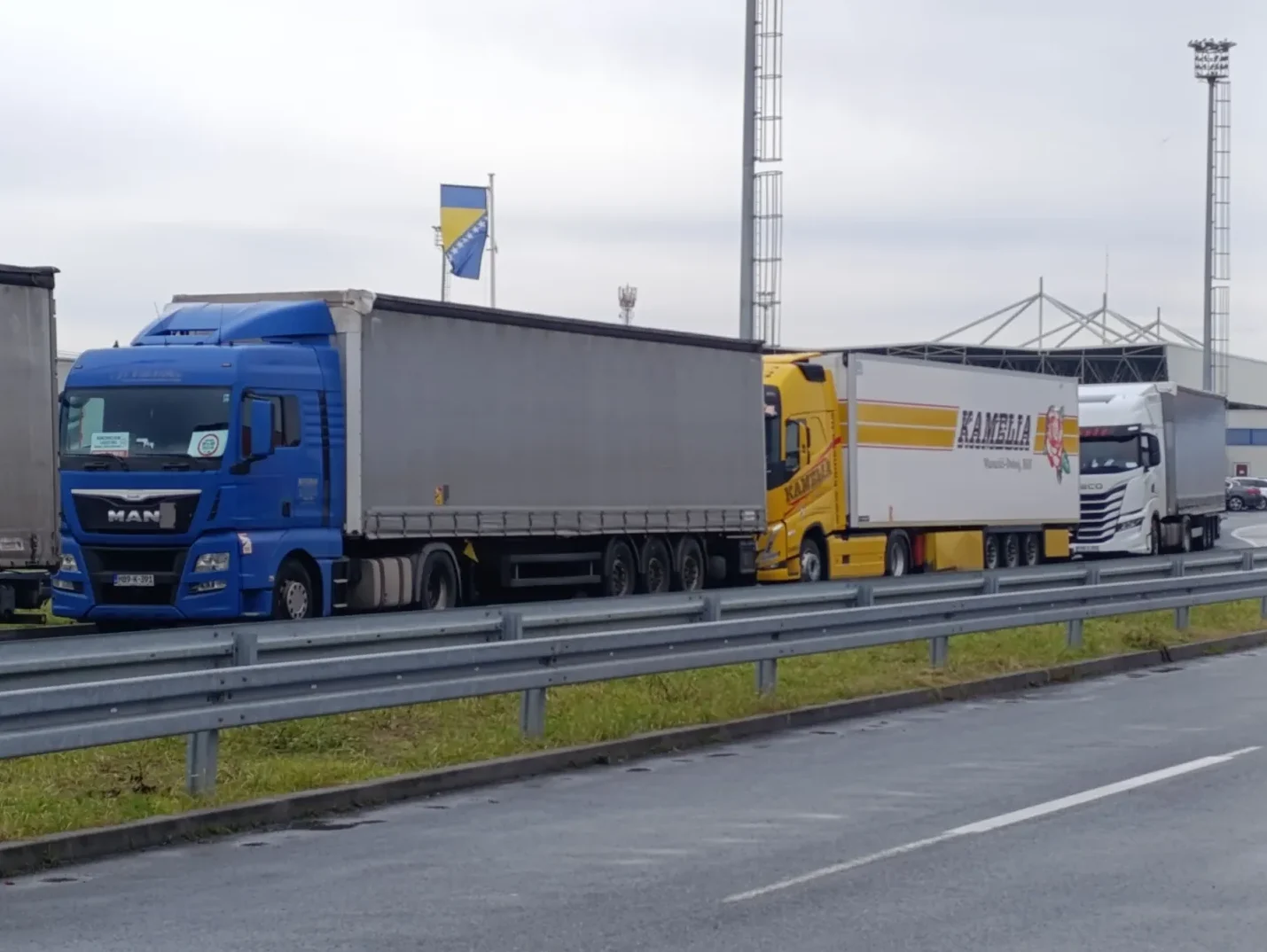 Kolona kamiona na graničnom prelazu, ističu se plavi kamion marke MAN, žuti kamion s natpisom "KAMELIA" i bijeli kamion marke IVECO.