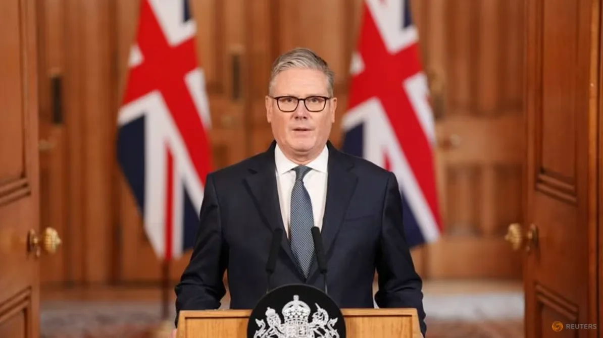 Keir Starmer, leader of the UK Labour Party, stands at a wooden podium with microphones in front of him. Two Union Jack flags are visible behind him, flanking him on either side. He is wearing a dark suit, a white shirt, and a patterned blue tie. He has glasses on and is looking directly at the camera.