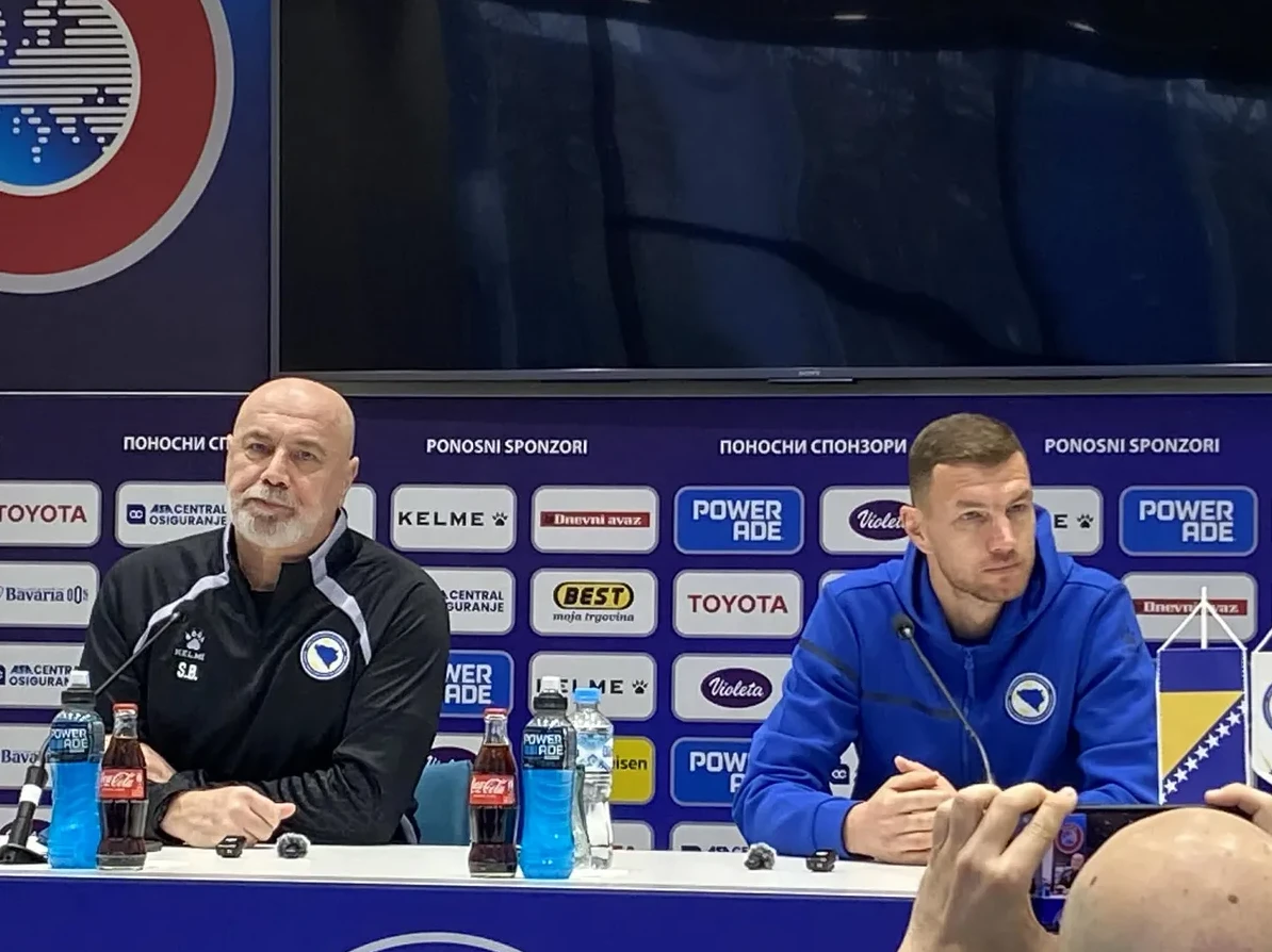 Dva muškarca sjede za stolom ispred natpisa sa sponzorima, dijeleći informacije na konferenciji za medije.