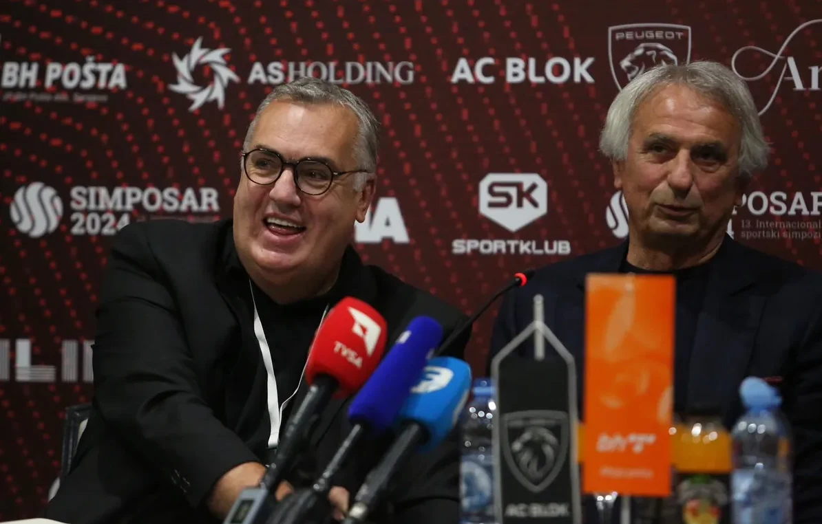 Dva muškarca sjede iza govornice sa logotipima sponzora, fokus je na muškarcu s naočarima koji se smije.