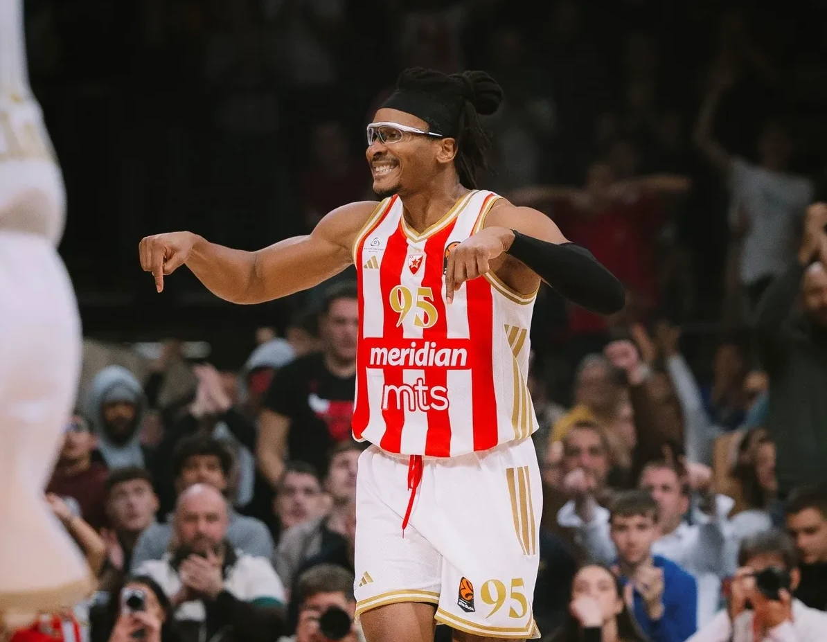Košarkaš sa naočarima i kosom vezanom u punđu, obučen u crveno-bijeli dres sa brojem 95 i natpisom "Meridian mts", gestikulira rukama prema dolje, dok publika u pozadini navija.