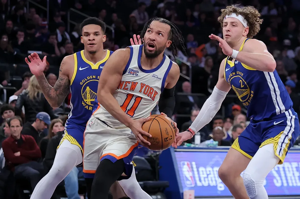 Košarkaš New York Knicksa s brojem 11, Jalen Brunson, u bijelom dresu sa natpisom "New York", drži loptu u rukama i gleda prema naprijed. S lijeve strane mu prilazi igrač Golden State Warriorsa u plavom dresu sa brojem 35, a s desne strane mu prilazi igrač Golden State Warriorsa s brojem 2. Obojica igrača Warriorsa nose bijele rukave na rukama.