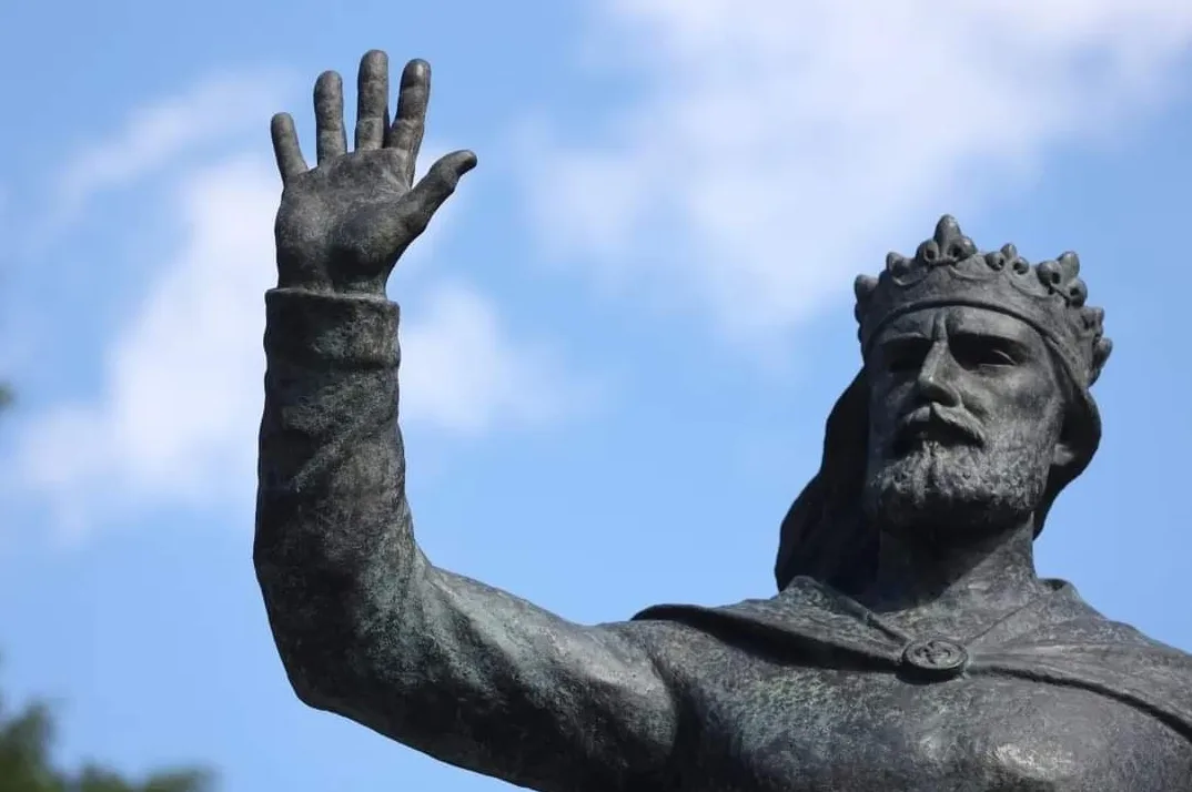 Bronzana statua kralja s krunom na glavi i podignutom desnom rukom u pozdravnom gestu.