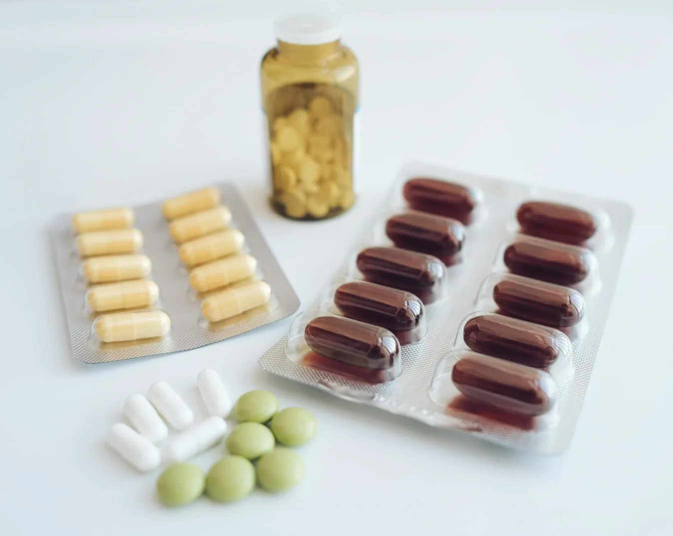 Blisteri sa žutim i smeđim kapsulama, nekoliko bijelih i zelenih tableta i smeđa boca s lijekovima na bijeloj podlozi.