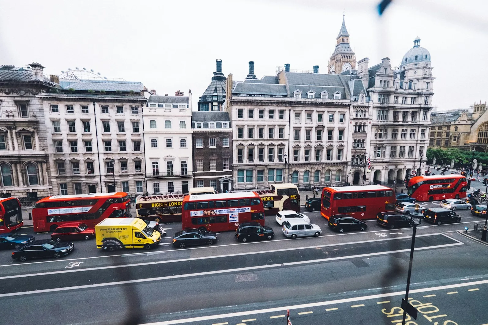 Crveni londonski dvospratni autobusi, žuta kombi vozila i automobili voze prometnom ulicom ispred starih zgrada u Londonu, sa Big Benom u pozadini.