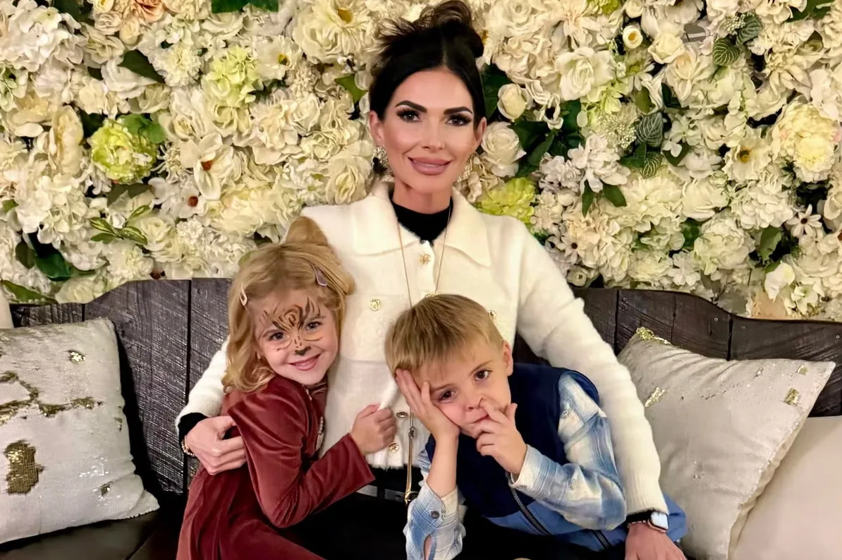 Majka sa dvoje djece pozira ispred cvjetne pozadine. Djevojčica ima oslikanu šminku na licu, a dječak je naslonio glavu na ruku.