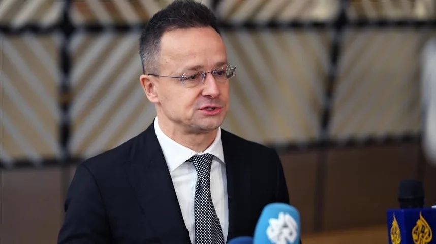 Peter Szijjarto, ministar vanjskih poslova Mađarske, nosi crno odijelo, bijelu košulju i crnu kravatu sa bijelim tačkicama. Ima naočare i gleda lijevo dok govori.