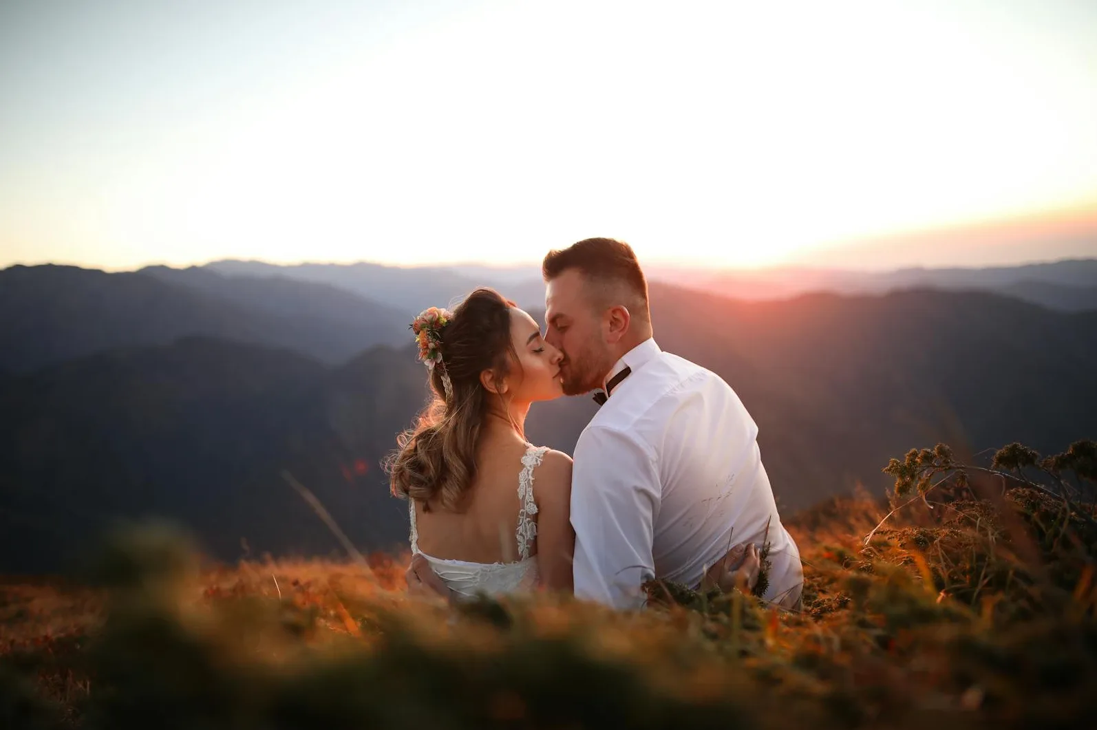 Mlada sa cvijećem u kosi i mladoženja u bijeloj košulji ljube se u zalasku sunca na vrhu planine, okruženi divljom travom.