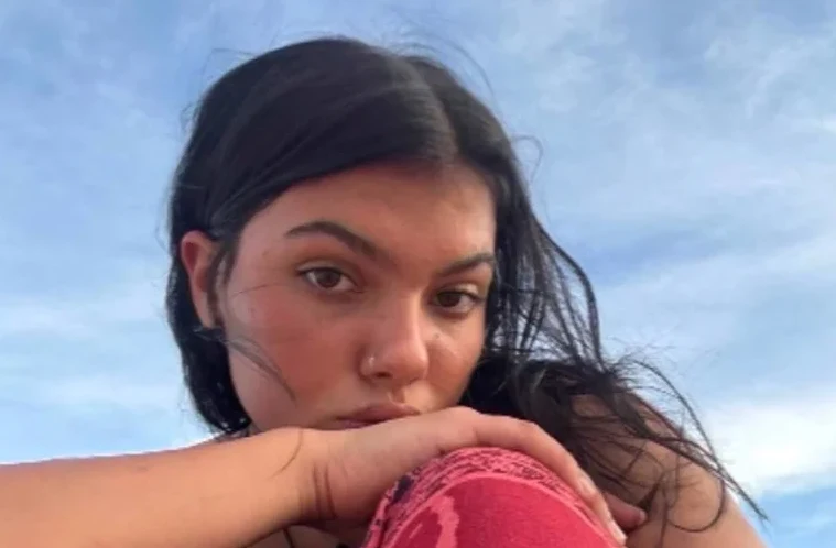 Portret mlade žene sa tamnom kosom, kako gleda u kameru, sa plavim nebom u pozadini.