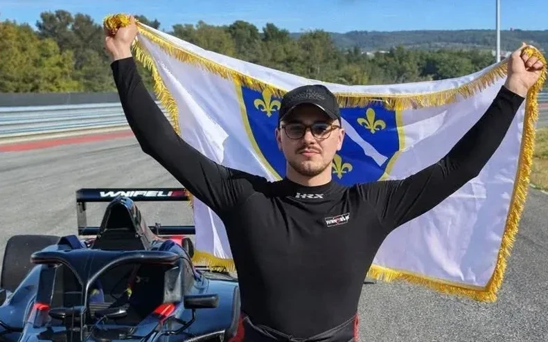 Mladi trkač u crnoj majici i crnoj kapi, sa naočalama, drži zastavu Kraljevine Bosne podignutu iznad glave. Na zastavi je prikazan dvoglavi orao sa ljiljanima na plavoj pozadini. Iza njega se vidi trkački automobil.