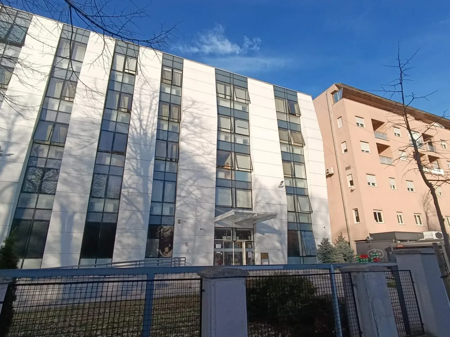 Savremena bijela zgrada sa puno staklenih prozora i nizom bijelih vertikalnih pruga, pored nje je narandžasta stambena zgrada, sve pod vedrim plavim nebom.