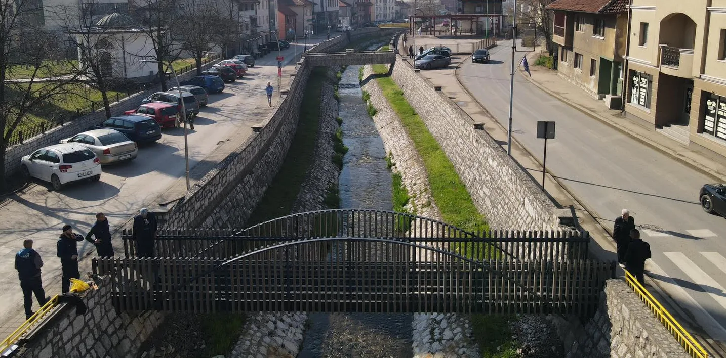 Most iznad gradske rijeke, okružen kamenim zidovima, sa parkiranim automobilima i pješacima.