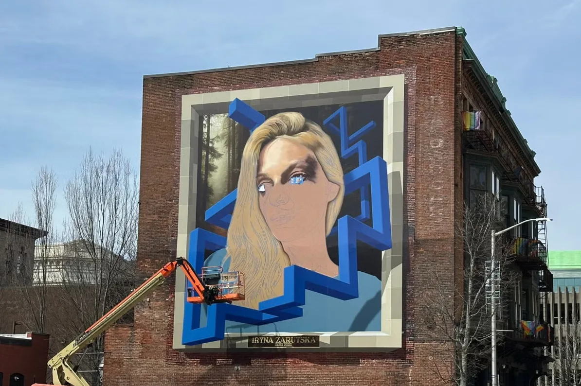 Veliki mural na ciglenoj zgradi prikazuje lice žene sa plavim elementima koji okružuju njenu glavu.