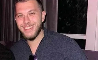 Mladić s kratkom smeđom kosom i bradom, nasmijan, nosi tamnu majicu na kopčanje.