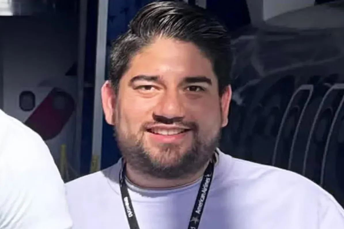 Mladić sa urednom bradicom i osmijehom na licu nosi ljubičastu majicu i lanyard "American Airlines". U pozadini se vidi dio aviona.