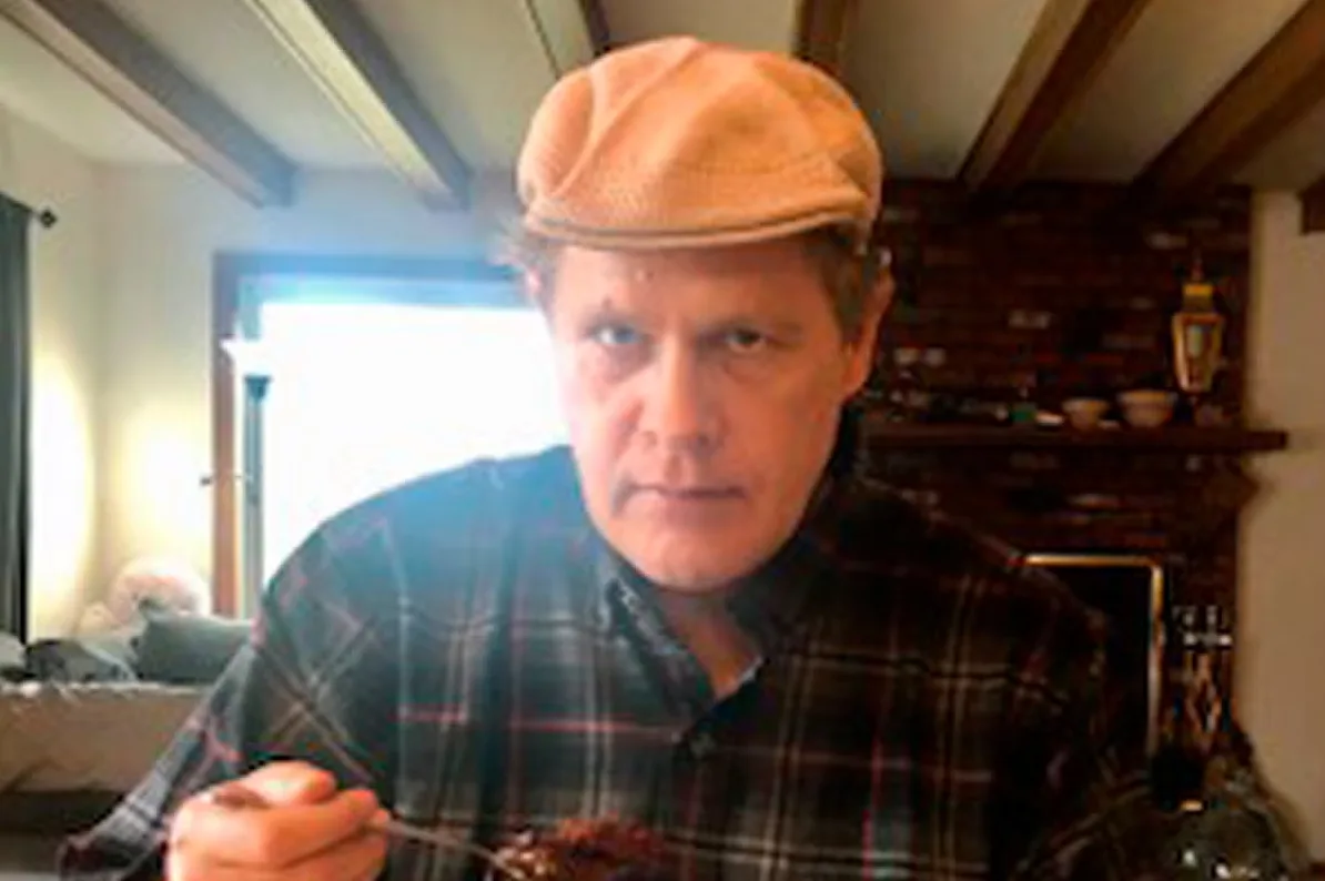 Muškarac u kariranoj košulji i bež kapi okreće se ka kameri dok jede iz duboke zdjele viljuškom.