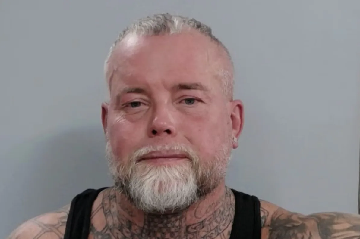 Portret muškarca sa sijedom bradom i kratkom kosom, obučen u crni prsluk, s vidljivim tetovažama na vratu i ramenima.