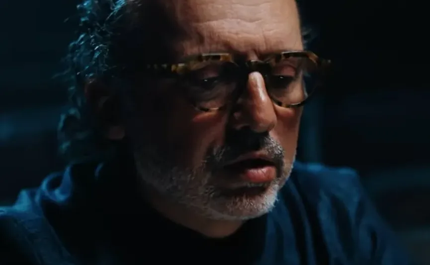 Muškarac srednjih godina sa naočarima i sijedom bradom, u tamnom okruženju