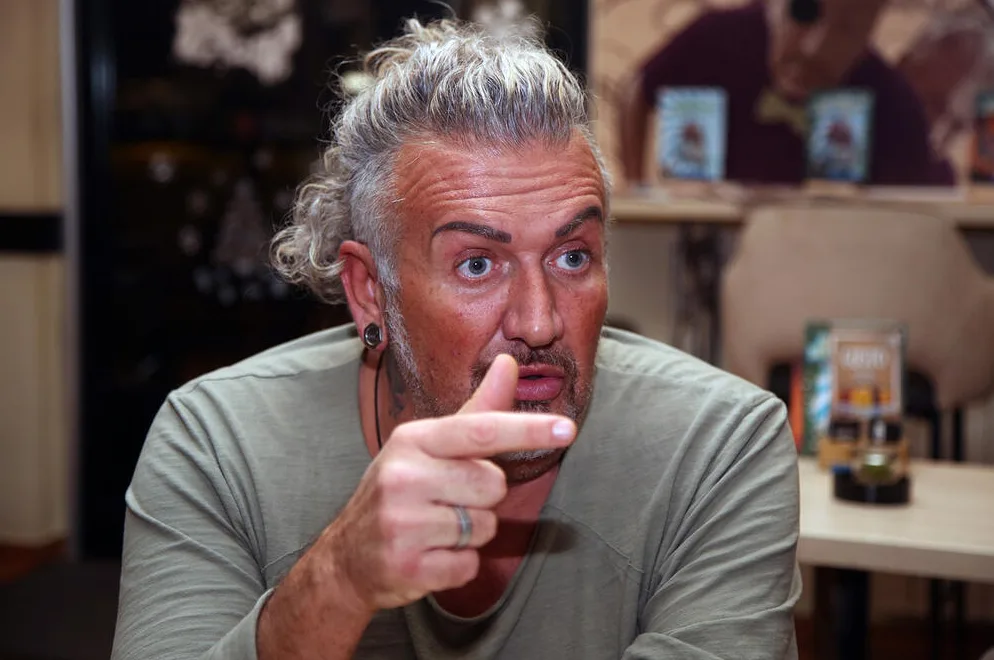 Muškarac sa neobičnom frizurom i brkovima, obučen u maslinasto zelenu majicu, pokazuje prstom u kameru, dok ga gleda sa iznenađenim izrazom lica.