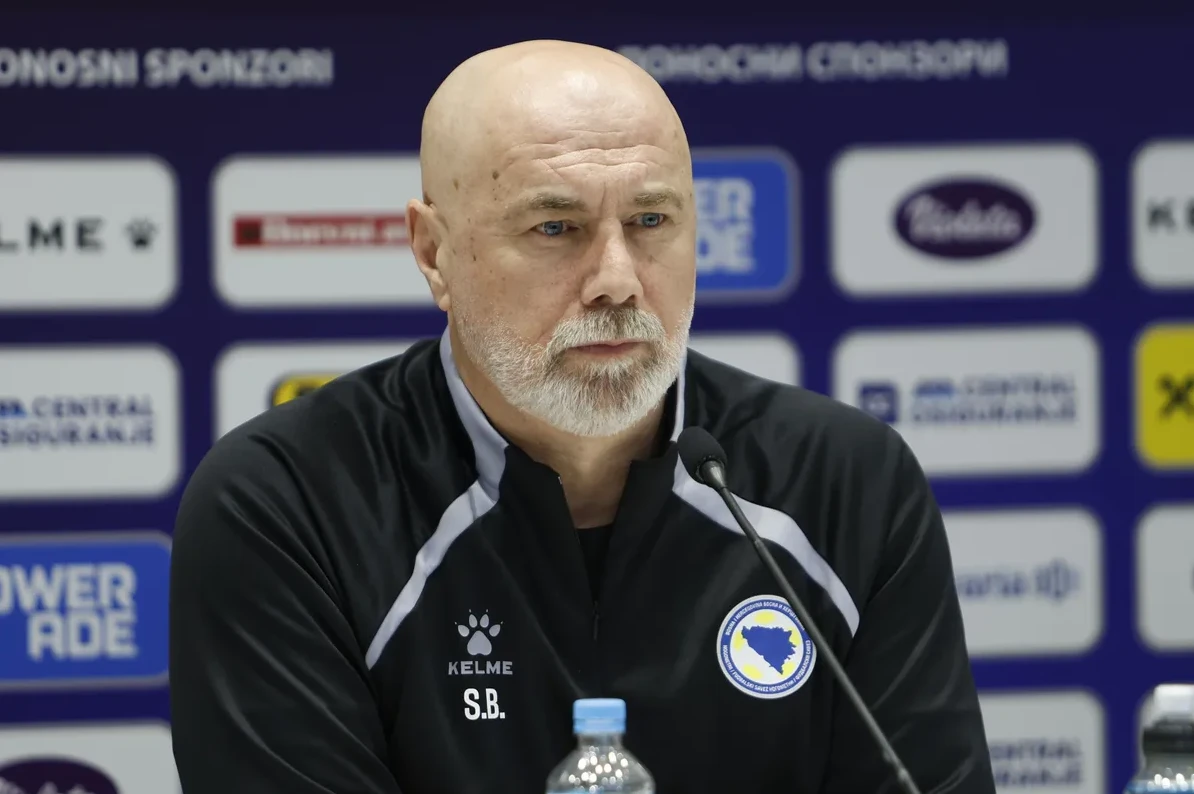 Ćelavi muškarac sa sijedom bradom, obučen u crnu sportsku jaknu sa grbom Bosne i Hercegovine, sjedi za govornicom