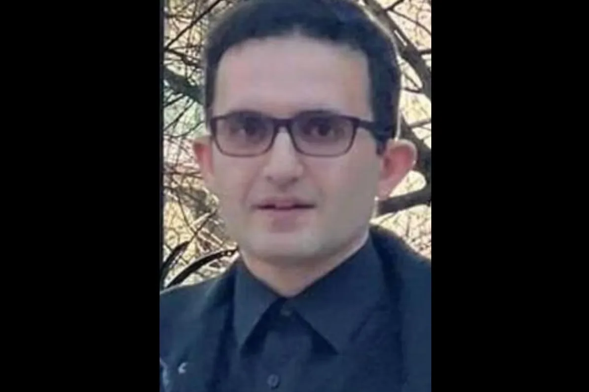 Portret mlađeg muškarca sa tamnom kosom, nosi crno odijelo i naočare, sa drvećem u pozadini.