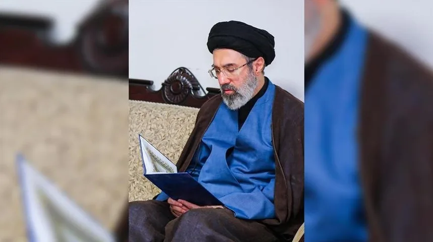 Muškarac u crnom turbanu i naočalama sa sjedinom bradom, obučen u plavi prsluk i smeđi ogrtač, sjedi na kauču i čita otvorenu knjigu.