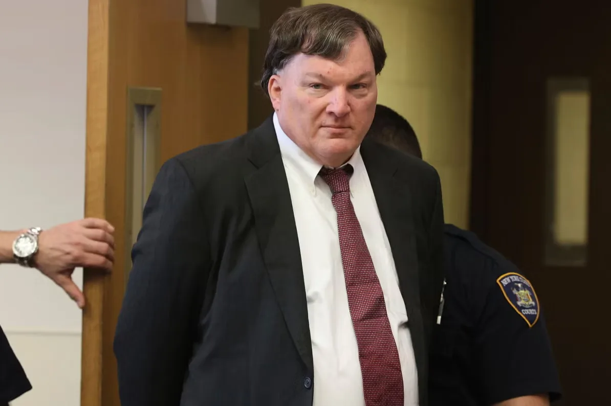 Muškarac u crnom odijelu sa bijelom košuljom i crvenom kravatom, gleda prema kameri.