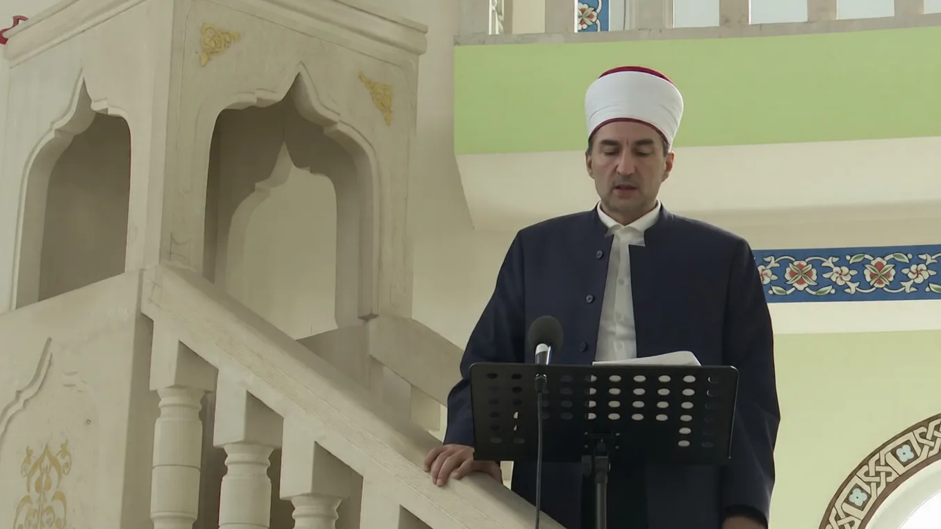 Imam u tamnom odijelu i bijelom turbanu čita sa stalka za note u džamiji.