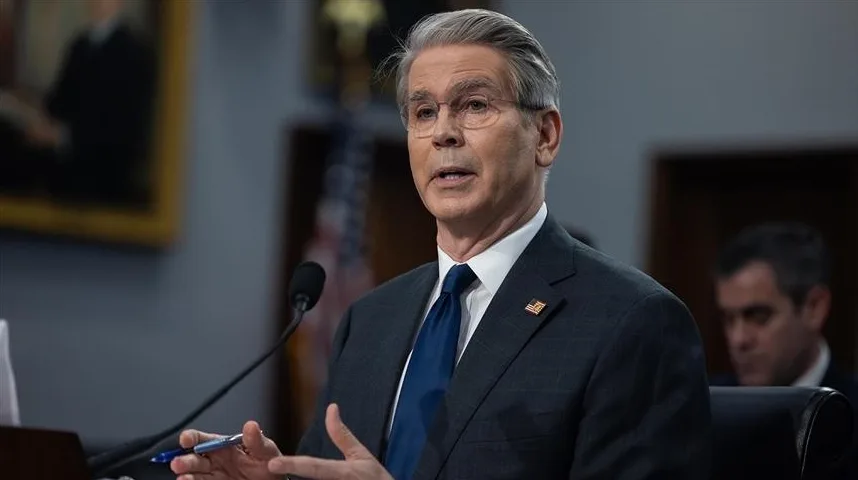 Muškarac u tamnom odijelu i plavoj kravati, sa naočalama, govori dok sjedi za stolom ispred mikrofona, držeći olovku u ruci.