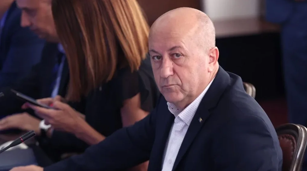 Muškarac u tamnom odijelu sa bijelom košuljom, sjedi za stolom, gleda u kameru.